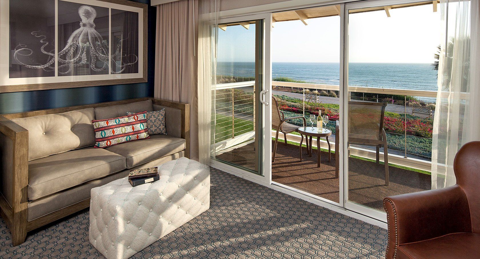 A cozy living room with a view: beige sofa, matching ottoman, artwork, and a sliding glass door opening to a balcony with ocean scenery, sunny and inviting.