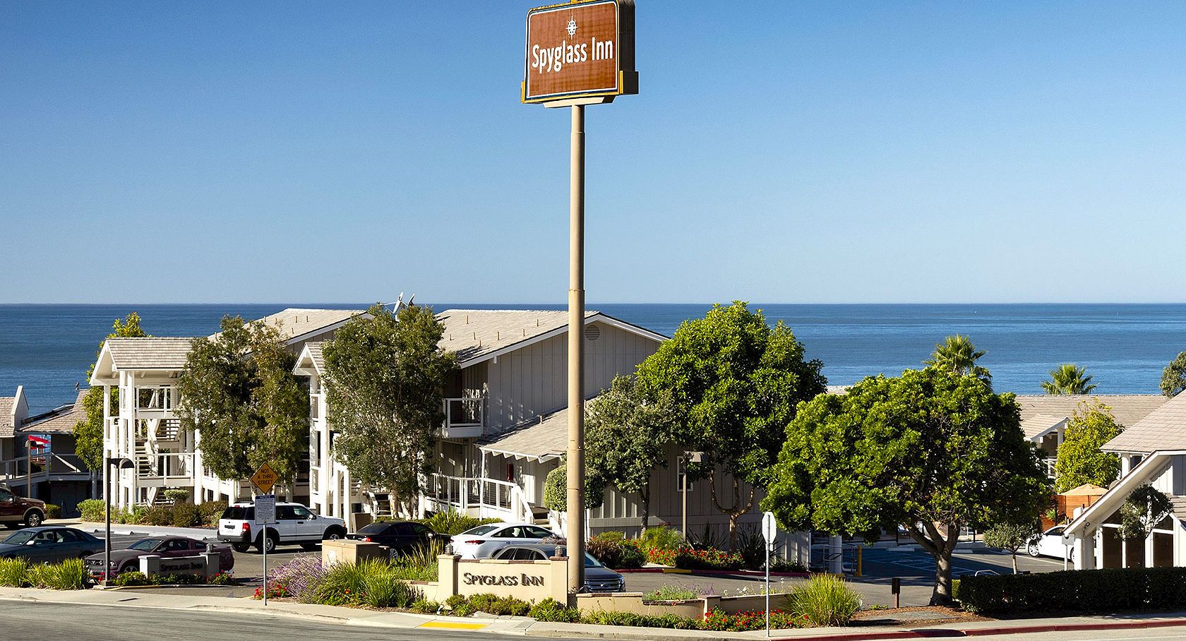 A small coastal motel on the shore, with a brown sign, trees, parking spots, and a view of the calm blue ocean under a clear sky.
