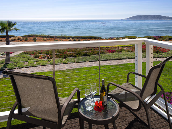 Two chairs and a small table on a balcony overlooking a grassy yard and ocean with a distant landmass in the background. The table holds a bottle, glass, and flowers.
