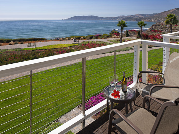 A balcony view overlooking a grassy lawn and ocean, with palm trees, flowers, and a small table with wine and glasses, perfect for sunset shots.