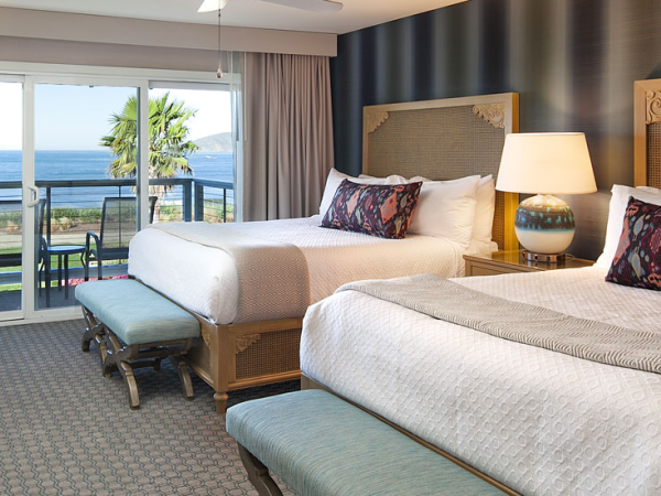A seaside hotel room with two queen beds, a balcony view of the ocean, matching lamps, decorative pillows, and a blue-accent wall.