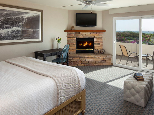 A cozy hotel room with a stone fireplace, mounted TV, desk, and a door to a balcony overlooking a sunny outdoor area, perfect for relaxing.