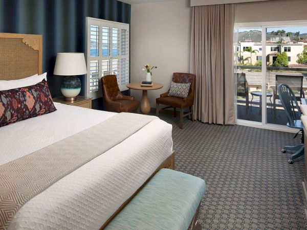 A hotel room with a large bed, seating area by a window, and a balcony view overlooking a sunny outdoor scene.