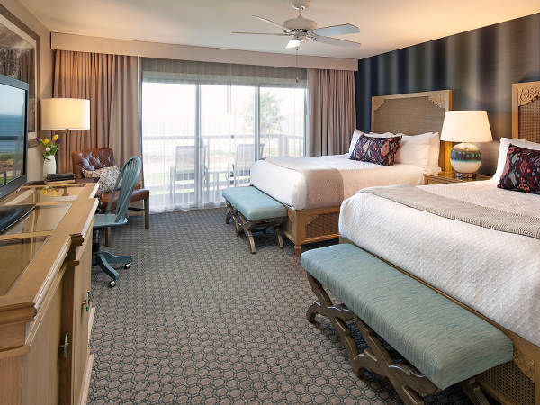 A hotel room with two queen beds, a desk and chair, a TV on a dresser, bedside lamps, a bench at foot of beds, and a balcony with sheer curtains.