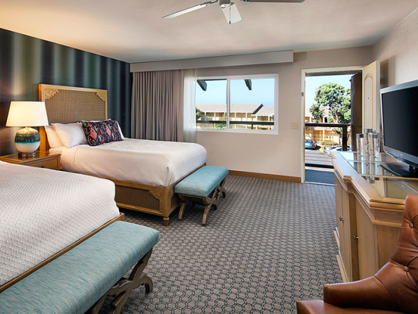 Two queen beds in a hotel room with a blue accent wall, nightstands, a desk, a TV, a lounge chair, and a balcony with a sunny view outside.