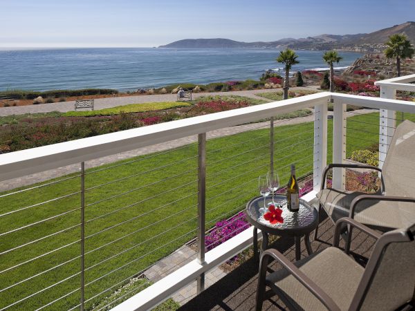 A seaside balcony with chairs, a small table with a bottle and glasses, and a view of the ocean, hills, and palm trees.