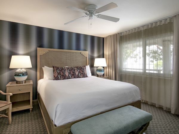 A hotel bedroom with a large bed, two nightstands and lamps, a blue accent wall, a window with sheer curtains, and an ottoman at the foot of the bed.