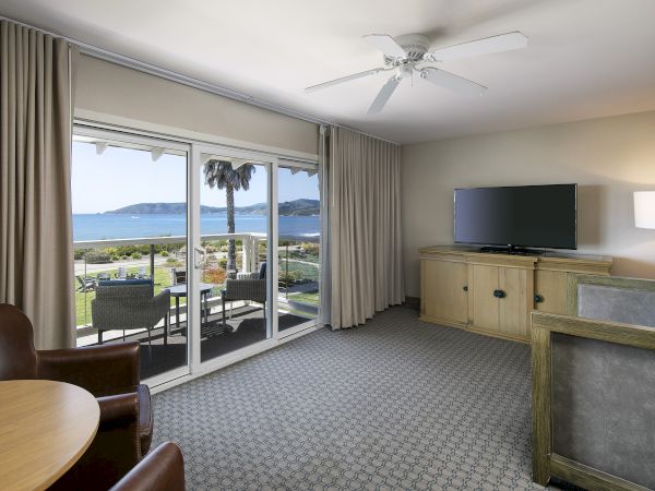 Cozy hotel suite with a sliding glass door to a balcony, a view of the ocean, beige carpet, wooden furniture, and a flat-screen TV, ending with a period.