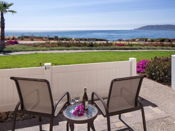A seaside patio with two chairs and a small table, overlooking a green lawn, pink flowers, and a calm blue ocean.