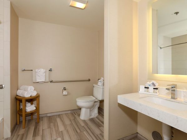 A modern bathroom with a toilet, bidet-style rails, a small wooden stool holding towels, a shower on the left, and a marble counter with a lit mirror on the right.