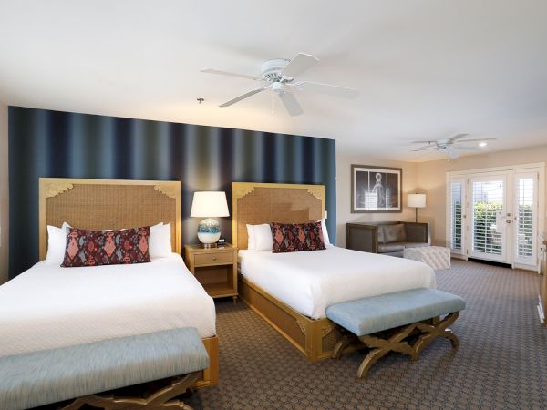 Two queen beds with padded headboards, a blue striped feature wall, matching benches, nightstands with lamps, and a seating/desk area by window doors.