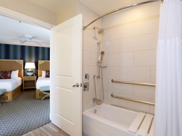 A hotel bathroom with a tub and shower, grab bars, and a doorway opening to a bedroom with two beds and lamps.