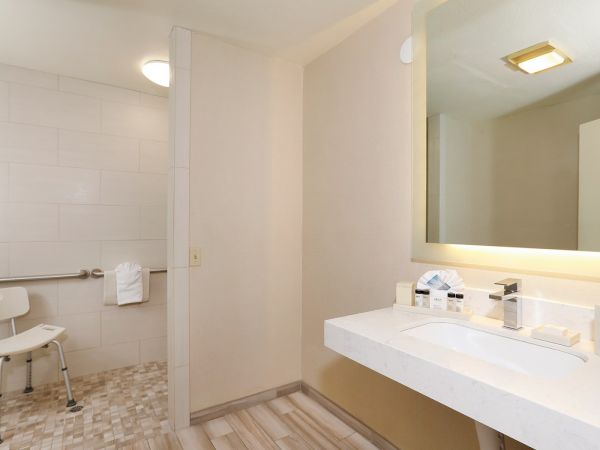 A clean hotel-style bathroom with a wide vanity mirror and sink on the right, a beige color scheme, and a private accessible-looking toilet and shower area to the left, with a chair in the shower and a towel rack.