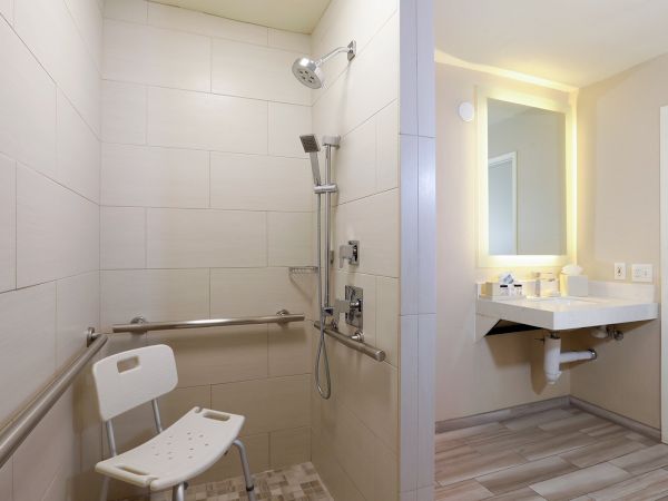 A accessible bathroom with a shower, grab bars, a foldable chair, and a separate vanity area with sink and mirror&mdash;bright and modern.
