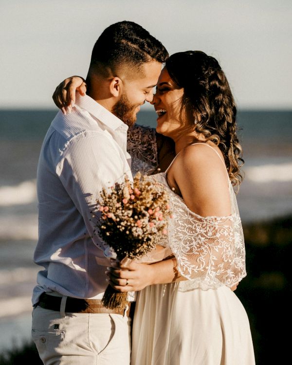 A couple stands close on the beach at sunset, smiling lovingly, embracing with a bouquet, sharing an intimate, joyful moment.