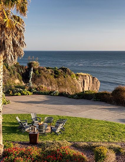 A sunny coastal scene with a palm tree, a grassy patio area, a few chairs around a table, and rocky cliffs meeting calm sea surf at sunset.