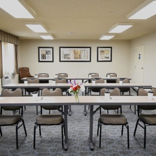 A small conference room with long tables and chairs arranged in rows, a flower centerpiece, wall art, and soft natural/fluorescent lighting.