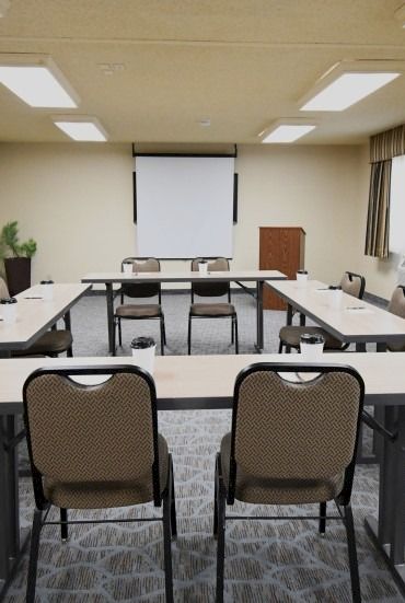 A conference room setup with a U-shaped arrangement of tables and chairs, a projector screen at the front, and a podium by the window.