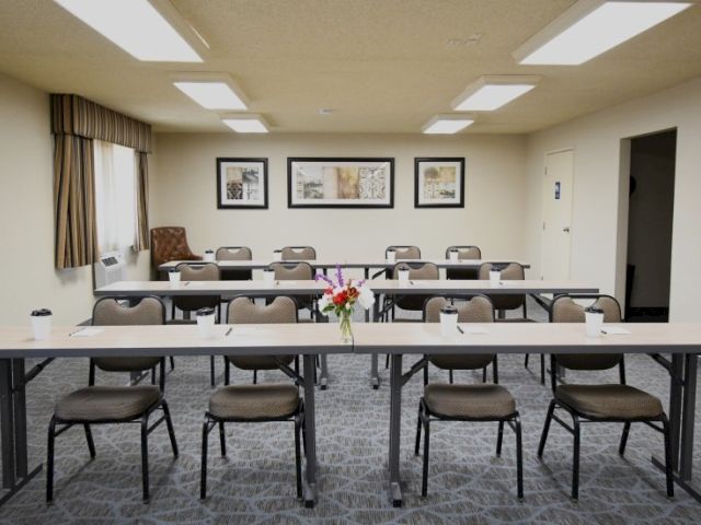 A small conference room with long tables, chairs, and a flower centerpiece; framed pictures on the back wall, lamps and windows on the left.