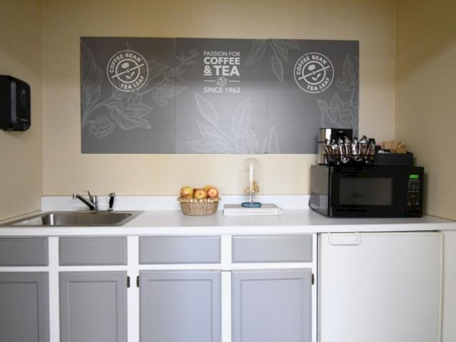 A small coffee/tea kitchenette with a chalkboard sign, sink, counter, basket of fruit, and a microwave on the right, plus white cabinets under.