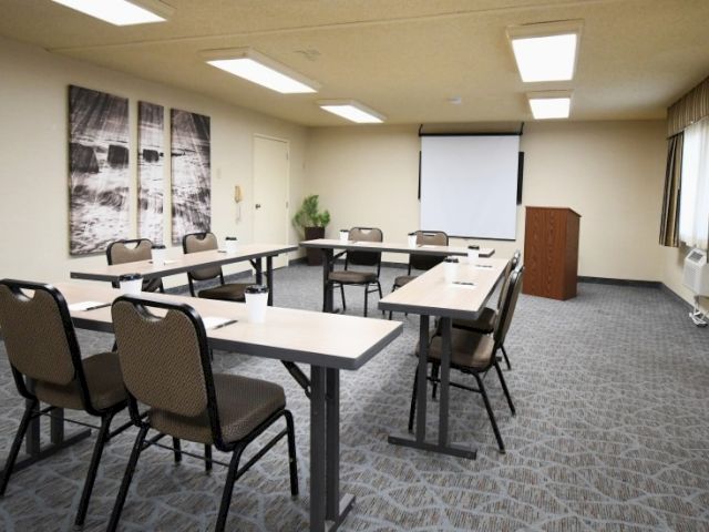 A small conference room with U-shaped desks, chairs, a projector screen, and neutral decor. End.