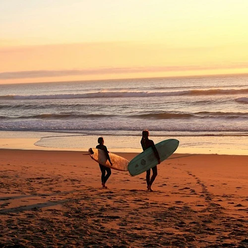 Two surfers walk toward the water with their boards at sunset on the beach.