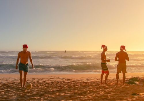 Three people enjoying a sunset on the beach; two chat near the water while one walks along the sand with a hat, calm waves in the background.