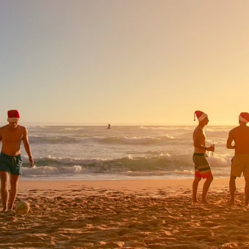 Three people enjoying a sunset on the beach; two chat near the water while one walks along the sand with a hat, calm waves in the background.