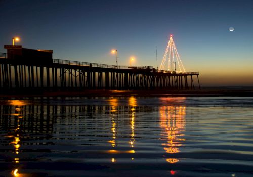 A waterfront pier at dusk with lights, calm water reflecting the glow, and a lit triangular structure or Christmas tree on the pier.