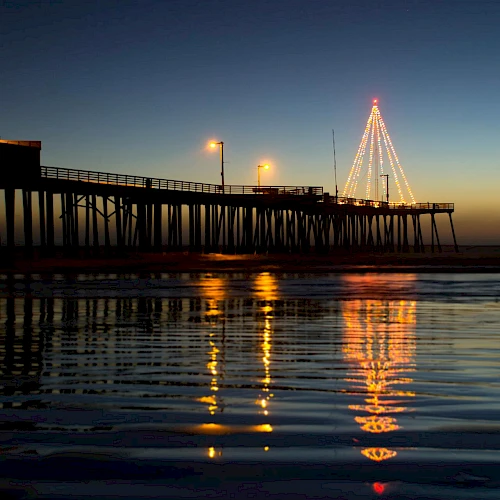 A waterfront pier at dusk with lights, calm water reflecting the glow, and a lit triangular structure or Christmas tree on the pier.
