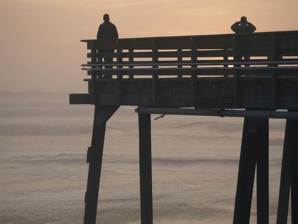A wooden pier extends into calm water at sunset; two silhouettes stand on the platform, watching the horizon as gentle waves glow.