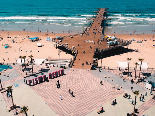 Aerial view of a bustling beach with a long wooden pier extending into the ocean; sunbathers, umbrellas, and palm trees fill a wide, tiled promenade.