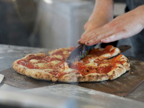 A chef slices a freshly baked pepperoni and cheese pizza on a metal board, steam rising as the knife cuts through the crust.