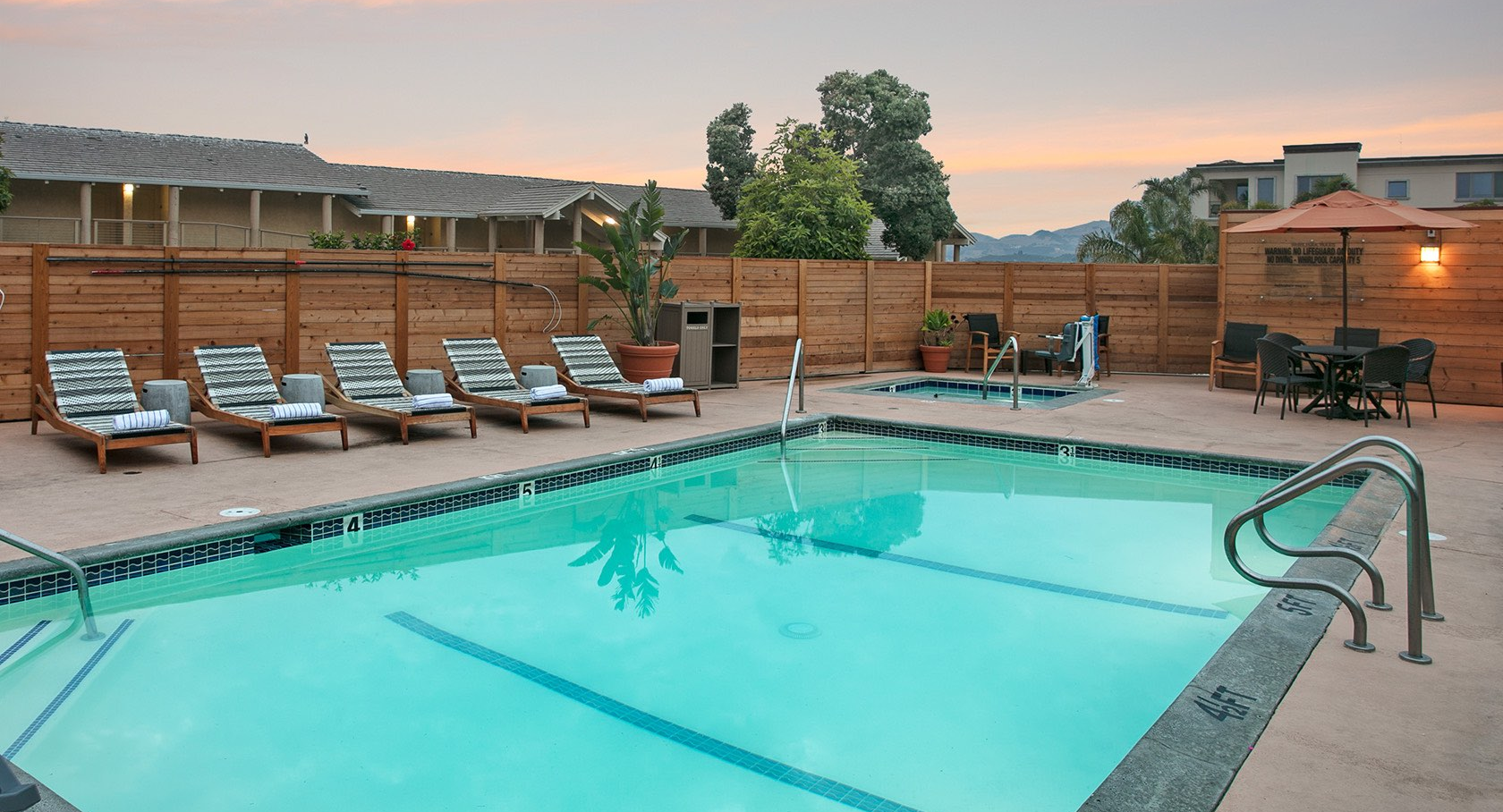 A clean outdoor pool area with turquoise water, sun loungers along the deck, a wooden fence, and a small dining setup in the corner at sunset.