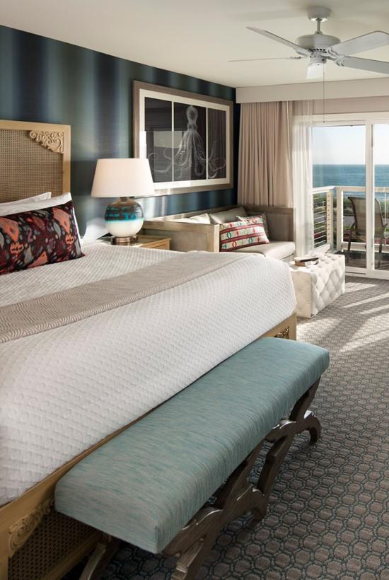 A hotel room with a king bed, teal bench, ample seating, desk, and a balcony view of the beach beyond sliding glass doors.