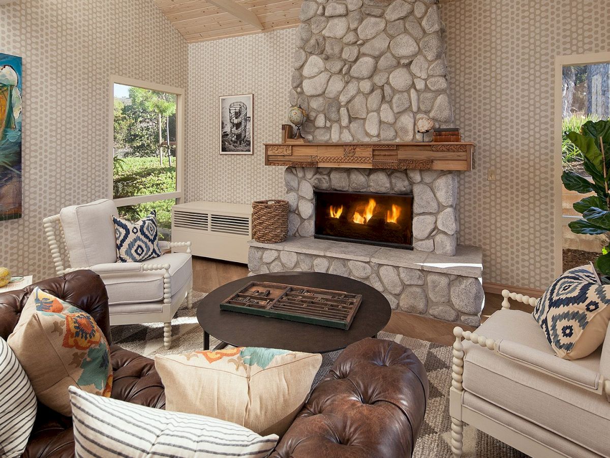 Cozy living room with a stone fireplace, leather sofa, and light cream chairs surrounding a glass coffee table, fire crackling warmly in the center.
