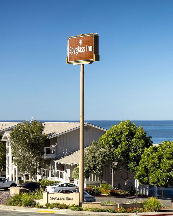 A seaside motel with a tall sign, beach vibe, trees, and a blue ocean backdrop; cars parked along the street by low-rise buildings.