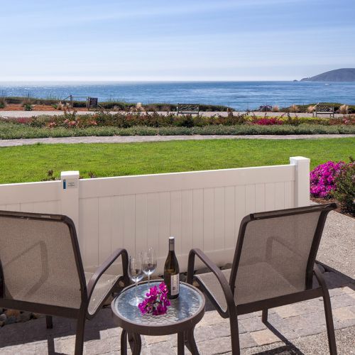 Two chairs and a small table on a patio overlook a grassy yard, ocean, and distant island; a bottle and flowers sit on the table.