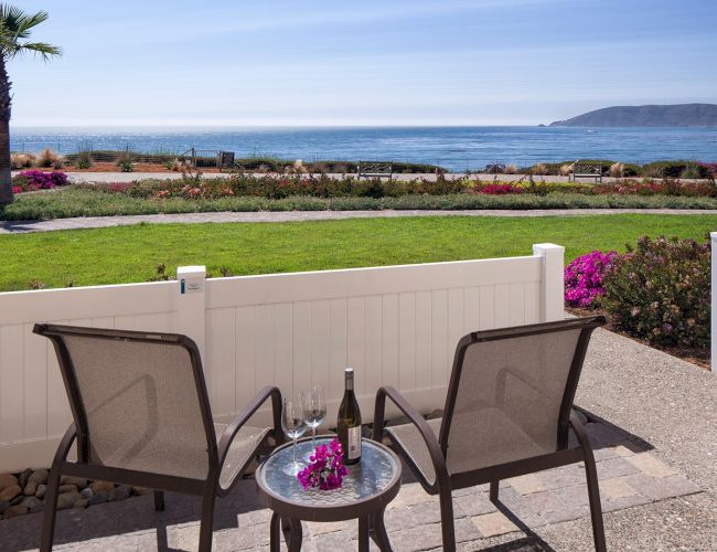 Two chairs and a small table on a patio overlook a grassy yard, ocean, and distant island; a bottle and flowers sit on the table.