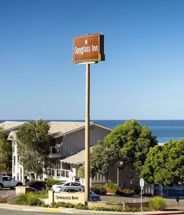 A roadside Inn near the beach with a tall brown sign, trees, parked cars, and a clear blue sky over the ocean.