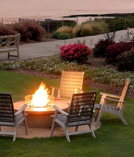 A coastal park at dusk with a lit fire pit surrounded by four chairs on green lawn, palm trees, and flowering shrubs along a winding path.