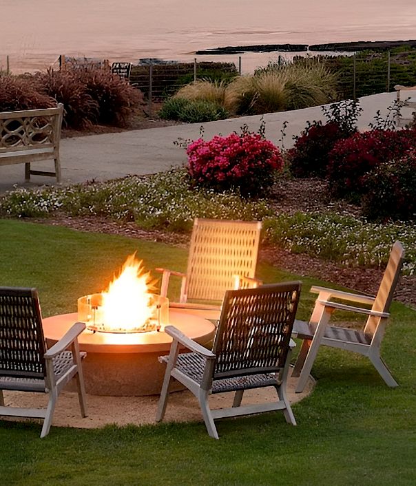 A coastal park at dusk with a lit fire pit surrounded by four chairs on green lawn, palm trees, and flowering shrubs along a winding path.