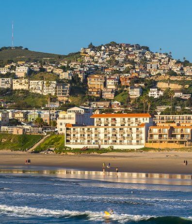 Coastal hillside town with colorful houses perched above a sunny beach and calm blue sea, waves gently rolling ashore along a long sandy shore.