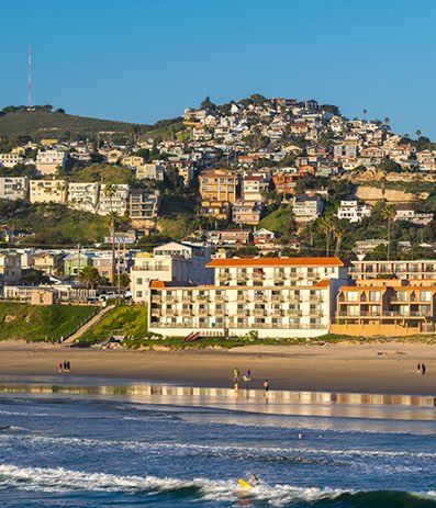 Coastal hillside town with colorful houses perched above a sunny beach and calm blue sea, waves gently rolling ashore along a long sandy shore.