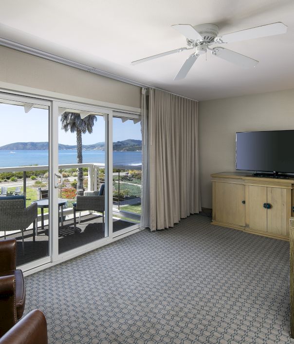 A hotel suite with a sliding glass door to a balcony, showing ocean views, beige carpet, a ceiling fan, a wooden console with a TV, and a sofa set, ending with the sentence.