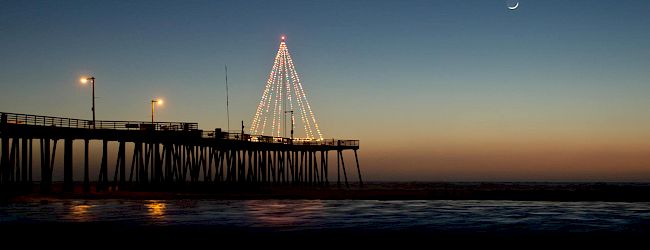 A pier at dusk with a lit Christmas-tree-shaped structure on stilts, calm water reflecting lights, and a crescent moon in the sky.