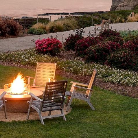 A seaside garden with a lit fire pit surrounded by four chairs, colorful flowers, palm trees, and a walkway along the coast at sunset, inviting and peaceful.