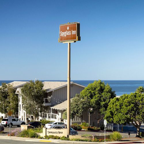 A small coastal motel by the beach with a brown sign, trees, and parked cars along a street near the ocean, under a clear blue sky.