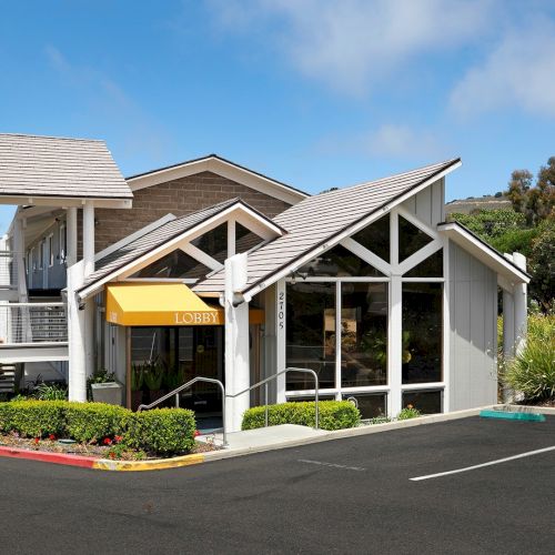 A modern small building with large glass windows, peaked white-trim roof, yellow awning, and a paved parking lot in front.