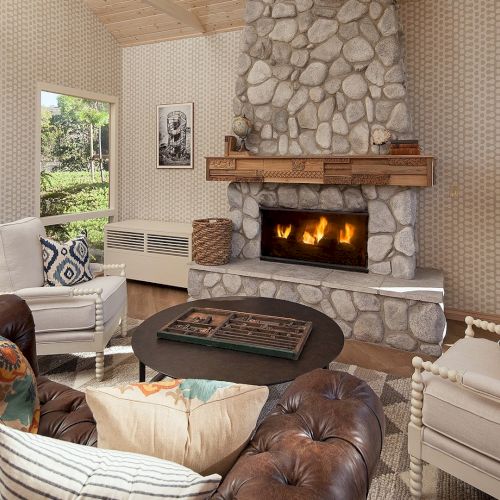 A cozy living room with a stone fireplace, leather and fabric sofas, throw pillows, a round coffee table, and plants by the windows.